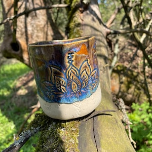Könnte beinhalten: Eine Keramiktasse mit weißer Glasur und einem blauen und braunen Blumendesign. Die Tasse steht auf einem mit Moos bewachsenen Baumstamm.