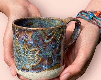 May include: A handcrafted ceramic mug with a distinctive floral pattern. The mug has a textured, glazed surface in hues of blue, brown, and gold. The handle is a matching brown. The base of the mug is natural, unglazed ceramic.