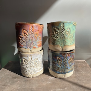 Może przedstawiać: Cztery ceramiczne kubki z motywem kwiatu lotosu. Kubki są ułożone parami, z dwoma górnymi kubkami w odcieniach brązu i zieleni oraz dwoma dolnymi kubkami w kolorze białym i niebieskim.