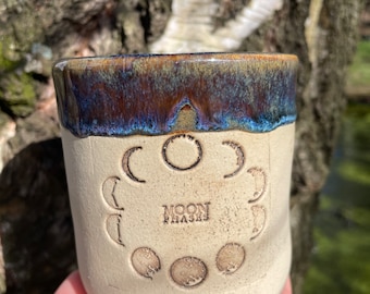 May include: A white ceramic mug with a blue and brown glaze. The mug has a design of the moon phases around the side and the words "Moon Phases" in the center.