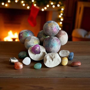 Può includere: Una collezione di bombe da bagno decorative con glitter iridescenti, disposte con varie pietre preziose lucide su una superficie di legno. Le bombe da bagno sono sferiche e le pietre preziose sono nelle tonalità del viola, verde, blu e arancione. Un camino e luci natalizie sono sullo sfondo.