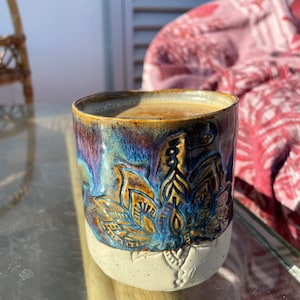 Puede incluir: Una taza de cerámica con un esmalte azul, marrón y blanco. La taza tiene un diseño floral y está llena de una bebida marrón claro.