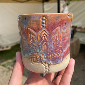 Könnte beinhalten: Eine Keramiktasse mit einem strukturierten, floralen Design in Braun-, Orange- und Blautönen. Die Tasse hat einen weißen Boden mit braunen Flecken.