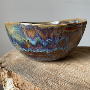 Puede incluir: Un cuenco de cerámica hecho a mano con un esmalte único. El cuenco presenta una mezcla de colores marrón, azul y dorado. El cuenco está colocado sobre una superficie de madera natural, mostrando su forma y textura orgánica. Este cuenco decorativo es ideal para servir o exhibir.