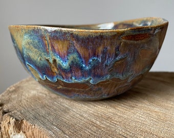 May include: A handmade ceramic bowl with a unique glaze. The bowl features a blend of brown, blue, and gold colors. The bowl is placed on a natural wood surface, showcasing its organic shape and texture. This decorative bowl is ideal for serving or display.