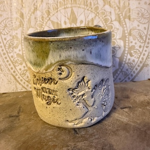 Könnte beinhalten: Eine handgefertigte Keramik-Tasse mit hellbeigem Äußeren und einem grün-blauen Rand. Die Tasse hat ein geprägtes Design einer Fee, eines Mondes und Sterne mit den Worten "Believe in the Magic".