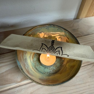 Könnte beinhalten: Eine Keramikschale mit türkisfarbener und goldener Glasur, die eine brennende Kerze enthält. Ein rechteckiges Keramikstück mit schwarzem Blumendesign liegt über der Schale. Die Flamme der Kerze beleuchtet das Innere der Schale.