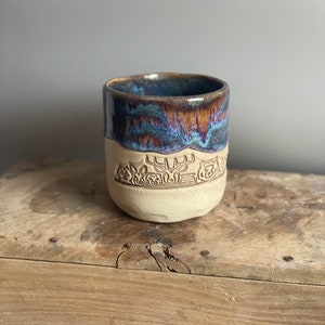 Może przedstawiać: Ceramiczny kubek z niebieską i brązową glazurą. Kubek ma biały pasek wokół środka z wypukłym wzorem jelenia i kwiatów.