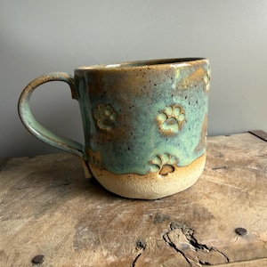 Op de afbeelding: Een keramische mok met een tweekleurig glazuur, met een gespikkeld blauwgroen en bruin bovengedeelte en een effen beige basis. Het buitenoppervlak is versierd met meerdere in reliëf aangebrachte pootafdrukken rondom de beker.