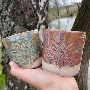 May include: Two ceramic mugs featuring a floral design. The mugs are glazed in a light brown and green colour with a white base. The floral design is a lotus flower.