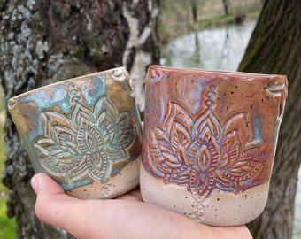May include: Two handmade ceramic cups with a lotus flower design. One cup is glazed in shades of blue and green, the other in brown and purple. Both cups have a speckled, off-white base and are held in a hand.