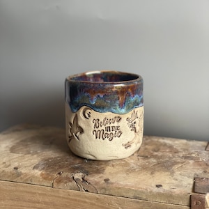 Puede incluir: Una taza de cerámica con un esmalte marrón, azul y morado. La taza tiene una inscripción blanca que dice "Cree en la magia".