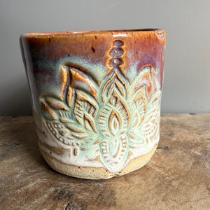 Könnte beinhalten: Ein Keramikbecher mit einem handgeschnitzten Lotusblumenmotiv auf der Vorderseite. Die Glasur verläuft von einem tiefen Rotbraun am Rand zu einem sanften, hellen Mintgrün über dem geprägten Blumenmuster, mit einer unglasierten, beigen Basis.