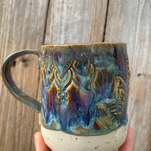 Könnte beinhalten: Eine handgefertigte Keramiktasse mit einer blauen, braunen und goldenen Glasur. Die Tasse hat ein strukturiertes, geprägtes Design und einen weißen Rand.