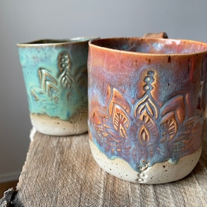 Könnte beinhalten: Zwei Keramikbecher mit floralem Design. Ein Becher ist türkis, der andere braun mit Blau- und Lilatönen. Beide Becher haben einen Henkel und einen hellen Boden. Die Becher stehen auf einer Holzoberfläche.