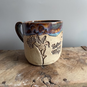 May include: A ceramic mug with a brown and blue glaze. The mug has a handle and a design of a fairy with wings and the words "Believe in the Magic" on the side.
