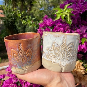 Puede incluir: Dos tazas de cerámica con un esmalte blanco y marrón. Ambas tazas presentan un diseño de mandala detallado. Una taza tiene un esmalte blanco con un diseño de mandala marrón, y la otra taza tiene un esmalte marrón con un diseño de mandala blanco.