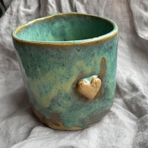 Puede incluir: Una taza de cerámica verde con un diseño en forma de corazón. La taza tiene un esmalte moteado y un borde marrón.
