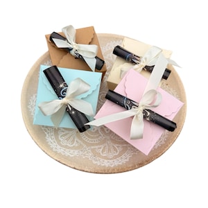 Könnte beinhalten: Vier kleine Geschenkboxen in Pastellfarben, jede mit einem schwarzen Stift und mit einem weißen Band gebunden, sind auf einem dekorativen Teller angeordnet. Die Boxen sind in Braun-, Creme-, Blau- und Rosatönen gehalten.