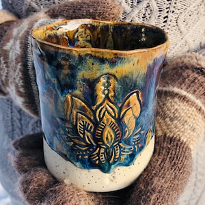 Op de afbeelding: Een keramische mok met een witte glazuur en een blauw en gouden bloemenontwerp. De mok is handgeschilderd en heeft een rustieke, handgemaakte uitstraling.