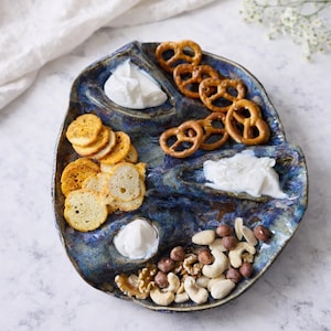 May include: A blue ceramic serving platter with multiple compartments, filled with pretzels, crackers, nuts, and dips. The platter has a unique, organic shape and a glossy finish, perfect for appetizers.