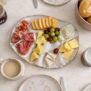 Könnte beinhalten: Ein rustikaler, cremefarbener Keramik-Servierteller mit einer Auswahl an Lebensmitteln, darunter Cracker, Käse, Trauben und Wurstwaren. Passende Teller mit Schneeflockenmustern, eine Brotschale und eine Kaffeetasse umgeben den Teller.