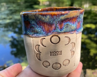 Könnte beinhalten: Ein Keramikbecher mit einer matten, cremeweißen Basis und einem glänzenden, irisierenden Rand in Blau-, Lila- und Bernsteintönen. Die Vorderseite zeigt ein geprägtes kreisförmiges Muster der Mondphasen um den Text "MOON PHASES" in Serifenschrift.