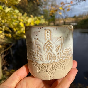 Puede incluir: Una taza de cerámica blanca con un esmalte marrón y un diseño floral blanco. La taza está salpicada de motas marrones.