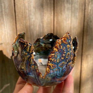May include: A small, ceramic candle holder with a unique, organic shape. The holder is glazed in a deep blue and green color with brown accents. The holder has a textured surface and appears to be handmade.