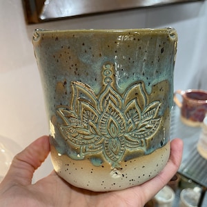 Könnte beinhalten: Handgefertigte Keramiktasse mit Lotusblumen-Design. Die Tasse hat eine zweifarbige Glasur, mit einem hellbeigen Sockel und einem teal-braunen oberen Bereich. Die Lotusblume ist in die Oberfläche der Tasse geschnitzt.