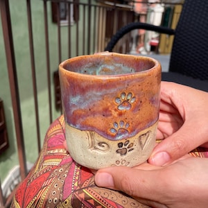Puede incluir: Una taza de cerámica hecha a mano con un diseño de huella de pata. La taza presenta un esmalte de dos tonos, con una parte superior marrón rojizo y púrpura y una base beige. La palabra "LOVE" y una huella de pata están talladas en la base.