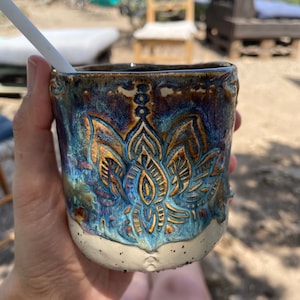 Könnte beinhalten: Eine blaue und braune Keramiktasse mit einem floralen Design. Die Tasse hat eine gesprenkelte Glasur und einen weißen Rand.