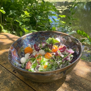 Könnte beinhalten: Eine Keramikschüssel mit einem bunten Salat aus orangefarbenen Kirschtomaten, Mozzarella-Kugeln, roten Zwiebeln und grünen Blättern. Die Schüssel hat eine dunkle, irisierende Glasur und steht auf einer rustikalen Holzoberfläche, mit einem Teich im Hintergrund.