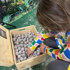 Könnte beinhalten: Eine Holzkiste gefüllt mit grauen, runden, glatten Steinen. Ein Kind greift in die Kiste, um einen Stein herauszunehmen. Die Kiste steht auf einer grünen Oberfläche, auf der graue Steine verstreut liegen.