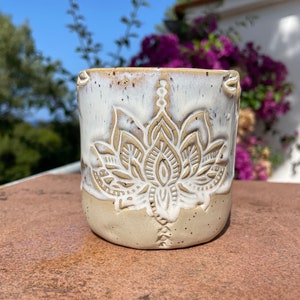 Puede incluir: Una taza de cerámica blanca con un esmalte marrón y un diseño de flor de loto blanca.