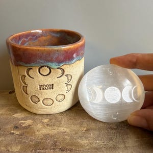 May include: A ceramic cup with a tan base and a reddish-brown glazed rim, featuring an embossed moon phase design and the text "MOON PHASES". Beside it is a smooth, translucent white stone disc engraved with a triple moon phase symbol.