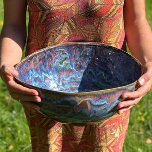 May include: A large, handmade ceramic bowl with a unique, iridescent glaze in shades of blue, purple, and bronze. The bowl has an organic shape and is held by a person. The bowl is suitable for serving or as a decorative piece.