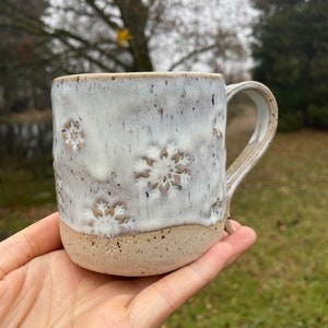 May include: A handmade ceramic mug with a white glaze and snowflake designs. The mug has a natural, unglazed base and a curved handle. The mug is held in a hand, showcasing its size and texture.