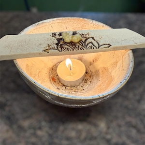 Könnte beinhalten: Eine Keramikschale mit einer brennenden Teelichtkerze im Inneren. Ein dekoratives Holzstück mit floralem Design und zwei kleinen, bernsteinfarbenen Objekten liegt über der Schale. Die Schale hat einen hellbeigen Innenraum und einen blau-grauen Rand.