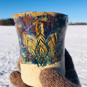 Könnte beinhalten: Eine Keramiktasse mit weißer Glasur und einem braunen, blauen und gelben Blumendesign. Die Tasse wird von einer behandschuhten Hand in einer schneebedeckten Landschaft gehalten.