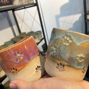 May include: Two ceramic mugs with paw print designs. One mug is a light brown with a dark brown paw print design. The other mug is a light blue with a light green paw print design.