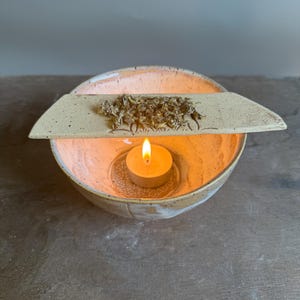 Könnte beinhalten: Eine gesprenkelte Keramikschale mit hellpfirsichfarbener Glasur, die ein brennendes Teelicht enthält. Ein flaches, rechteckiges Keramikstück mit strukturierter Oberfläche und getrockneten Kräutern liegt auf der Schale und zeigt ein geometrisches Muster.