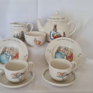 May include: A vintage cream-colored tea set with rabbit illustrations and text. The set includes a teapot, creamer, bowl, two plates, and two teacups with saucers. The plates and teapot have the text "Once upon a time there" and "into mischief, I am".