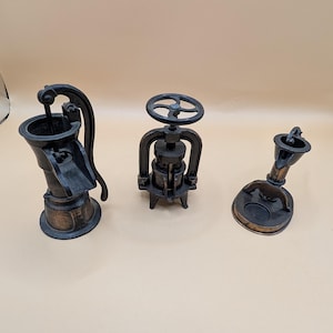May include: Three miniature metal objects with a dark bronze finish. On the left, a hand-operated water pump. In the center, a press with a circular top wheel. On the right, a small funnel-shaped device with a circular base and a side spout.