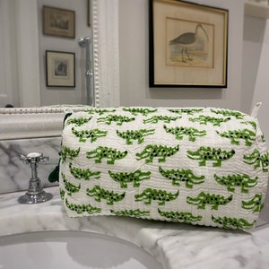 May include: White quilted makeup bag with a green alligator print. The bag is sitting on a white marble countertop.