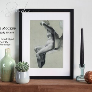 May include: A black framed mockup with a drawing of a nude figure sitting on a stool. The frame is on a wooden shelf with a green glass vase, a small potted succulent, a black candle, and a glass candle holder with a brown orb.