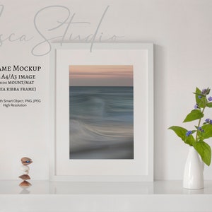 May include: White frame mockup with a seascape photograph inside. The frame is sitting on a white shelf with a small white vase with purple flowers on the right side.