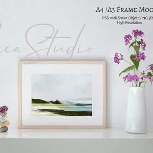 May include: A white frame mockup with a light blue and green landscape painting inside. The frame is sitting on a white shelf with a vase of purple flowers and a stack of stones.