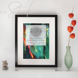 May include: A black framed art print mockup with a white mount, displaying a vibrant abstract painting. The frame holds an A4 image. A sprig of orange flowers in a green vase and a stack of stones are on the white surface.