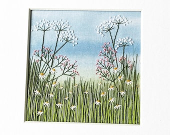 Peut inclure: Une broderie encadrée représentant un champ de fleurs sauvages blanches avec des accents roses sur un fond de ciel bleu.